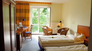 Habitación de hotel con dos camas, un escritorio con silla, una ventana con cortinas y vistas a zonas verdes.