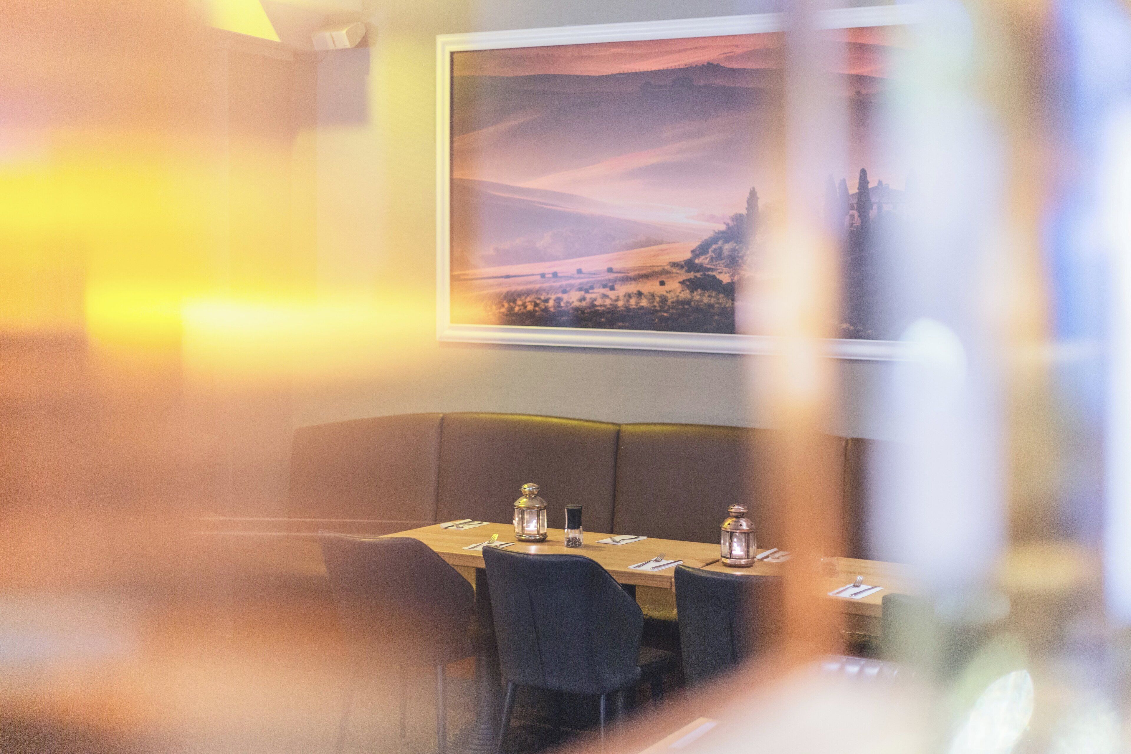 Un restaurante con una mesa dispuesta para dos, un cuadro de paisaje enmarcado en la pared y una iluminación ambiental cálida.
