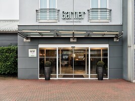 Exterior del hotel Garnier, un IHG Hotel, con una fachada moderna, una gran entrada de vidrio y plantas en macetas a la entrada.