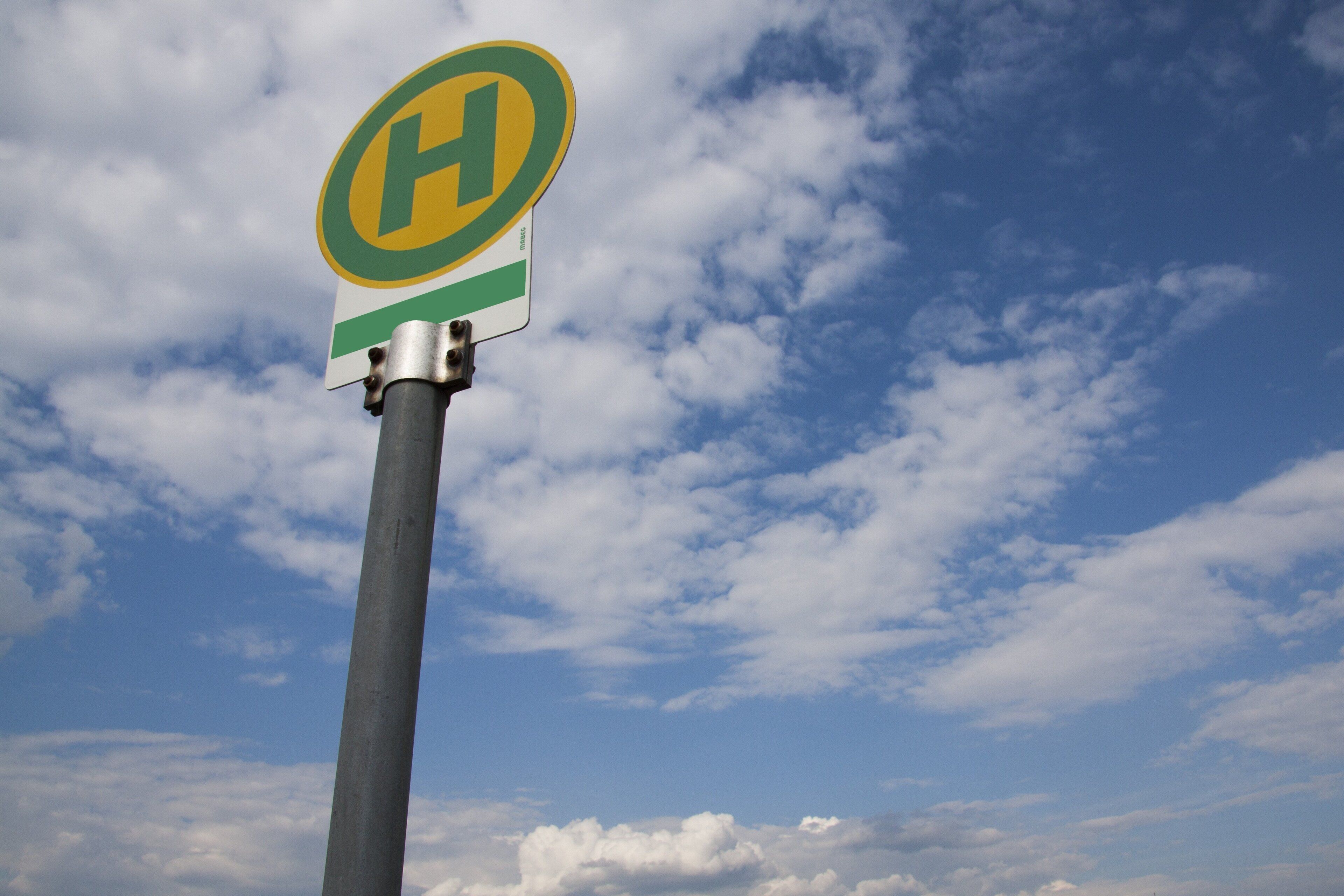 A sign with a green and yellow 'H' on a pole against a sky with clouds.