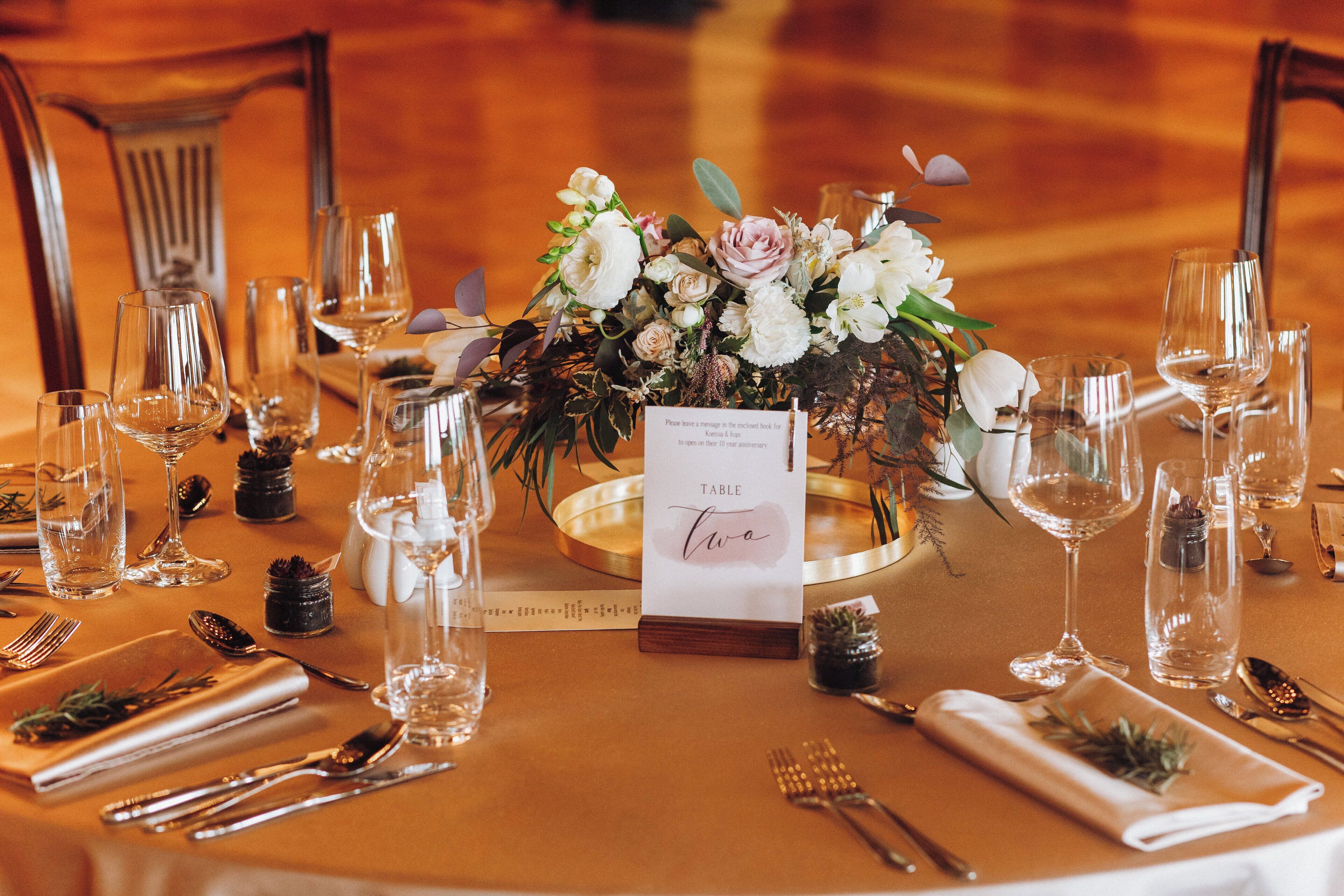 Una mesa de comedor elegantemente dispuesta, con un centro de flores, un letrero con el número de la mesa y cubertería ordenada.