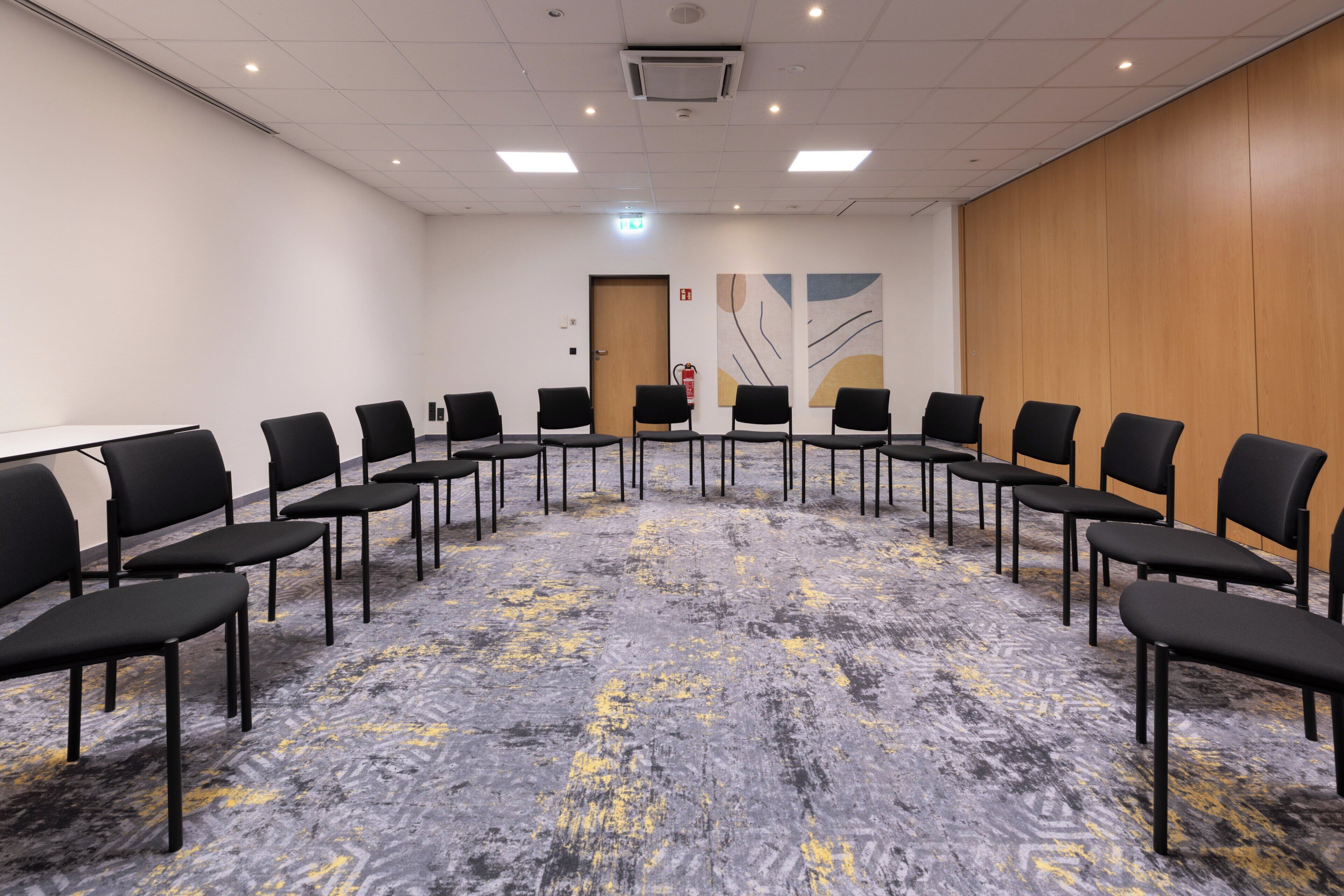 A conference room with a carpeted floor, black chairs arranged in rows, a wooden panel wall, and a door with a fire extinguisher.