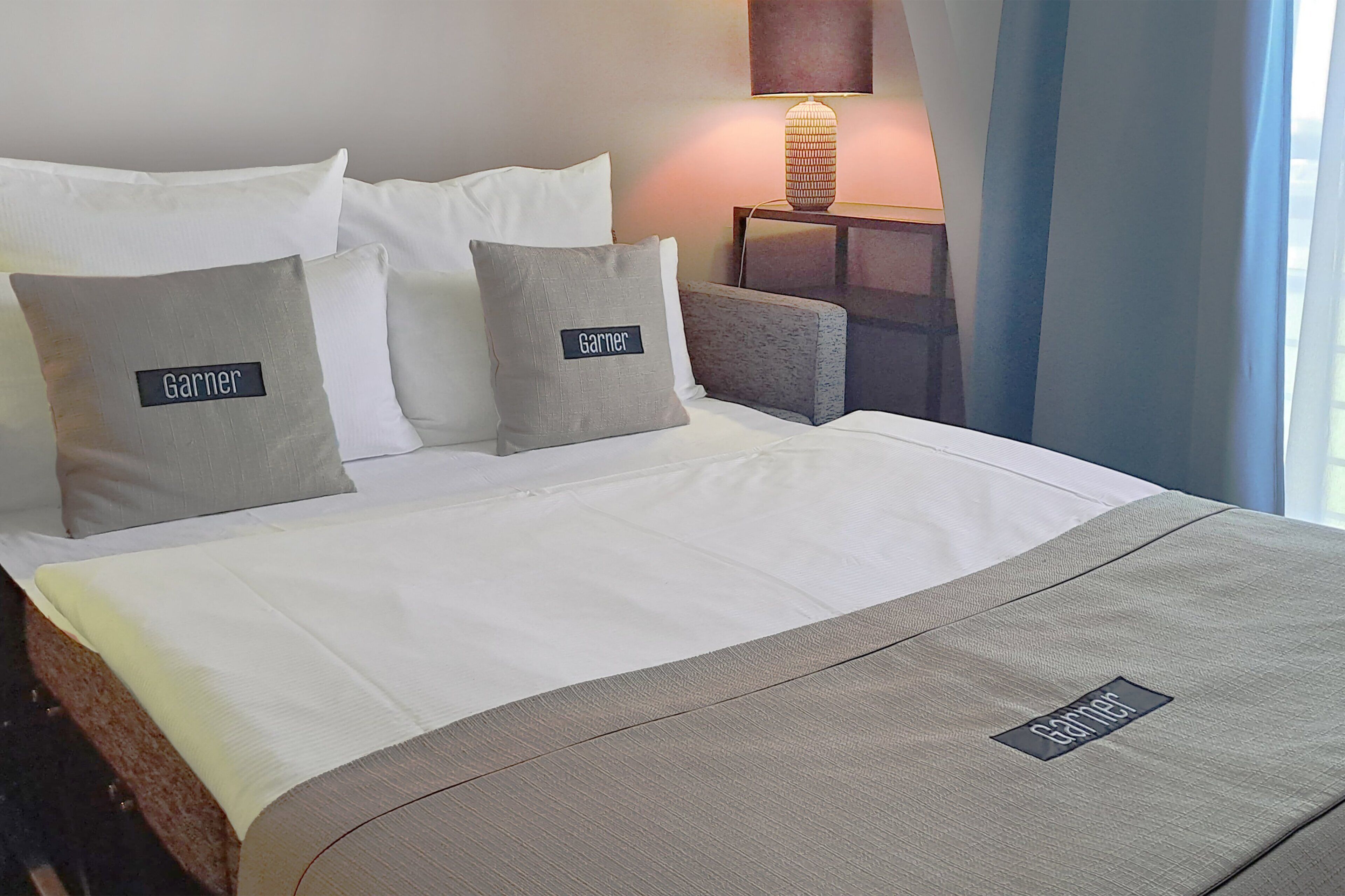 Una cama bien hecha con ropa de cama blanca y gris, dos almohadas con el nombre "Garnier" y una mesita de noche con una lámpara.