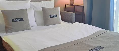 Una cama bien hecha con ropa de cama blanca y gris, dos almohadas con el nombre "Garnier" y una mesita de noche con una lámpara.