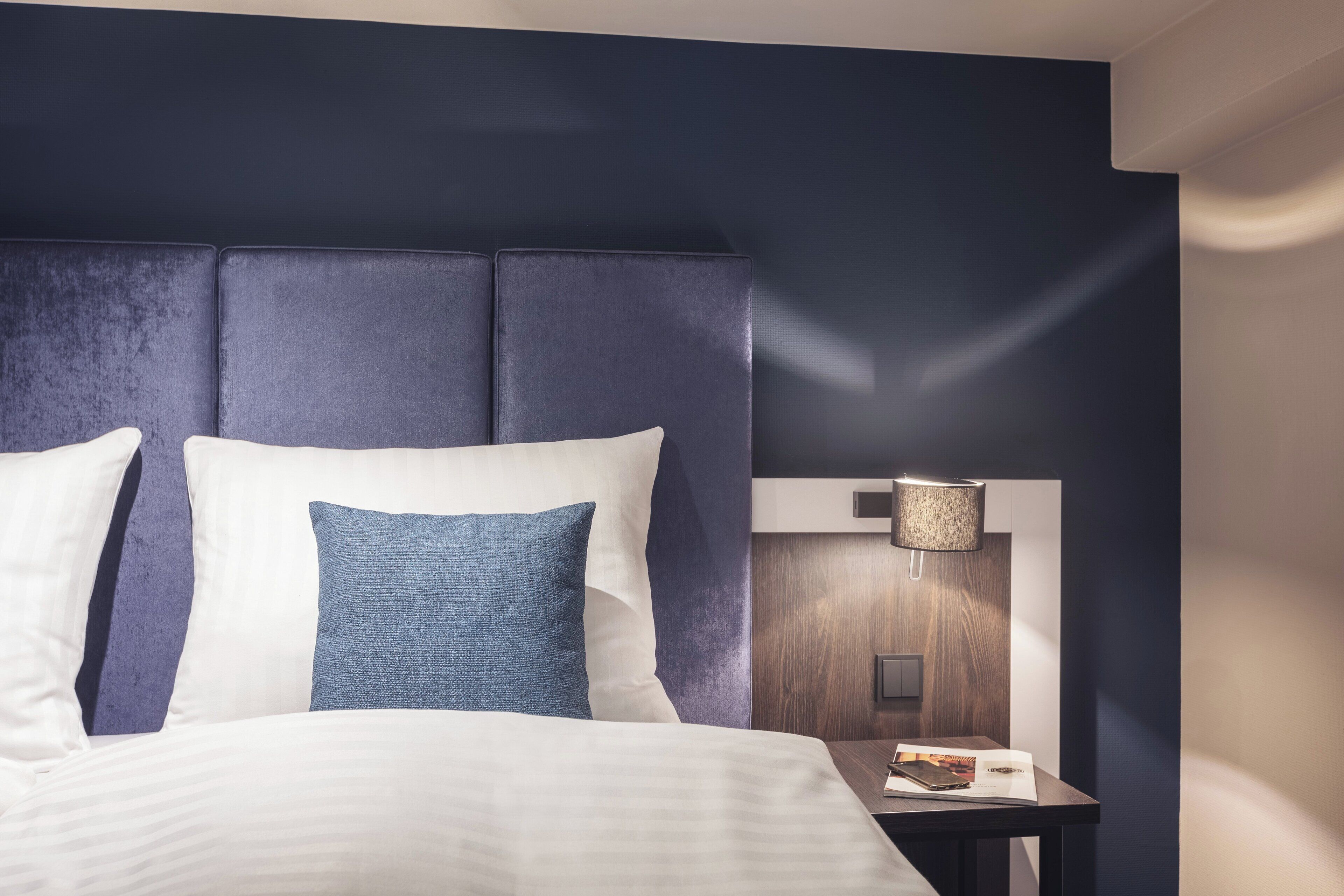 Habitación de hotel con una cama grande, una mesita de noche con lámpara y un cabecero oscuro.