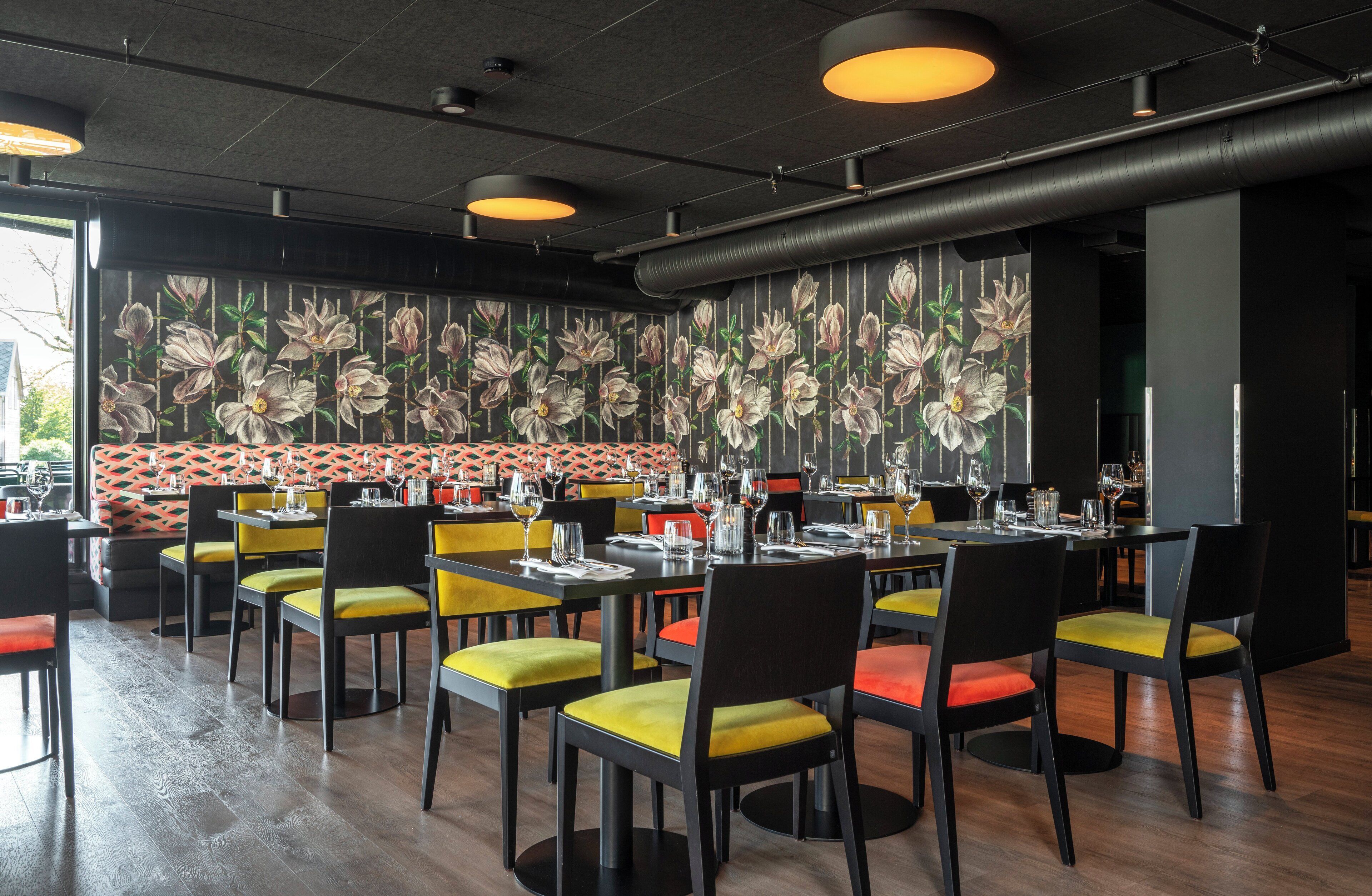 A restaurant interior with tables set for dining, chairs with yellow and red cushions, and a floral wall mural.