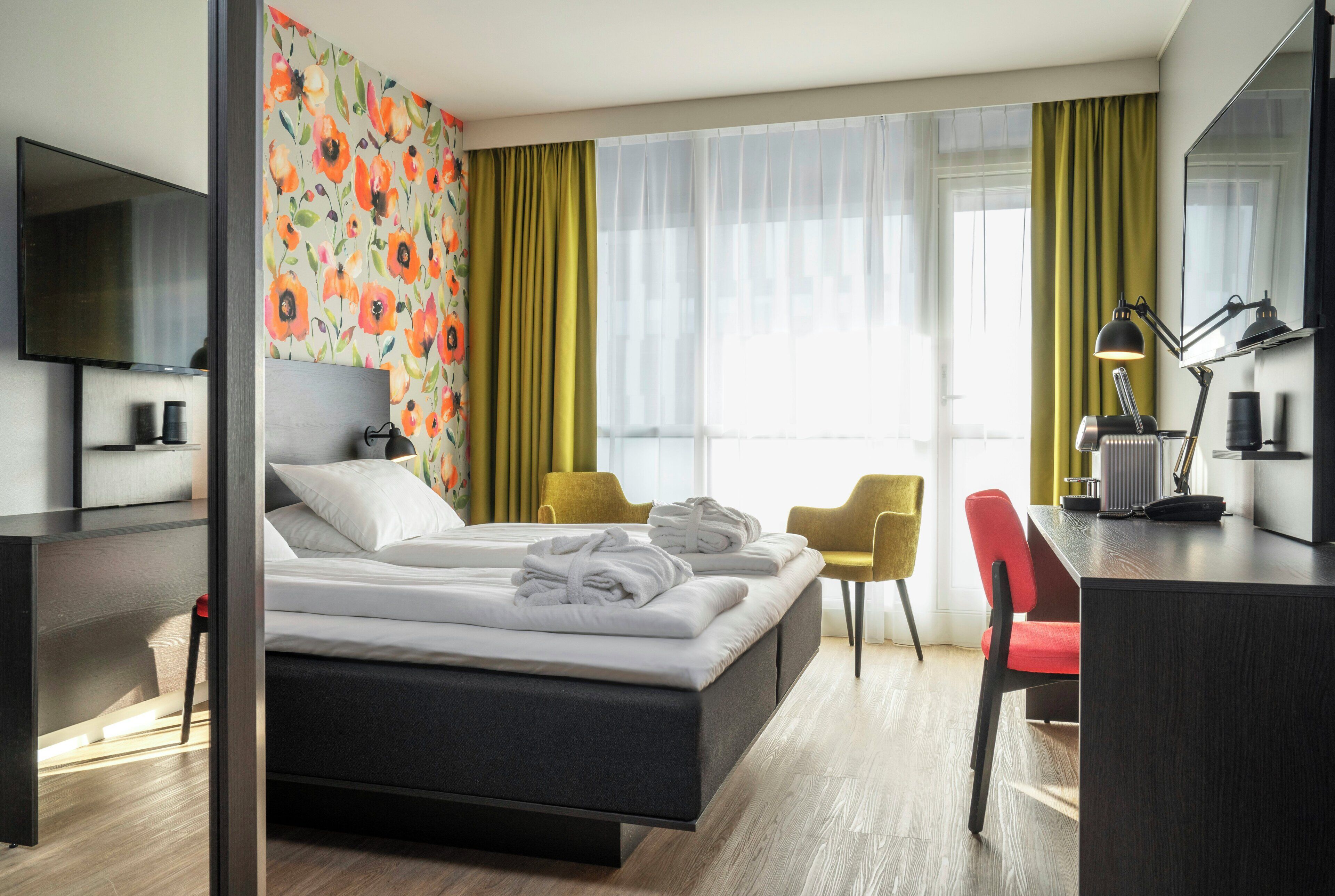 Habitació d'hotel moderna amb un llit, una taula de treball, una cadira, un televisor i una finestra gran amb cortines.