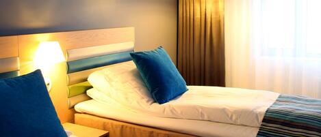 A hotel room with two beds, each with a headboard, a bedside table, and a lamp. The room has a window with curtains and a view of the outside.