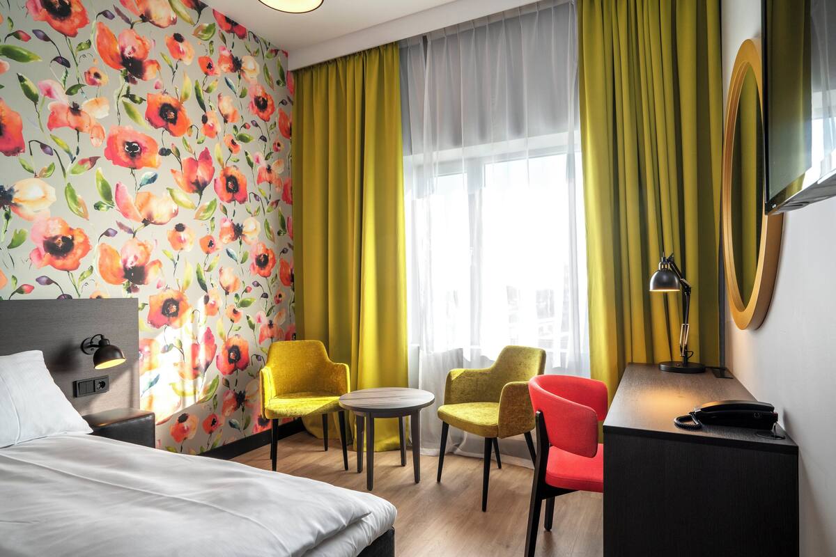 A hotel room with a bed, two yellow chairs, a red chair, a small table, a desk with a lamp, and a large window with curtains.