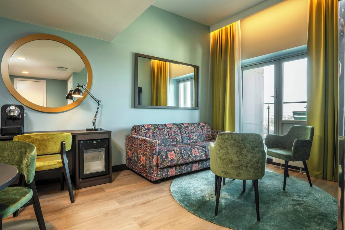 A modern living room with a sofa, chairs, a round mirror, a small heater, and a large window with curtains.
