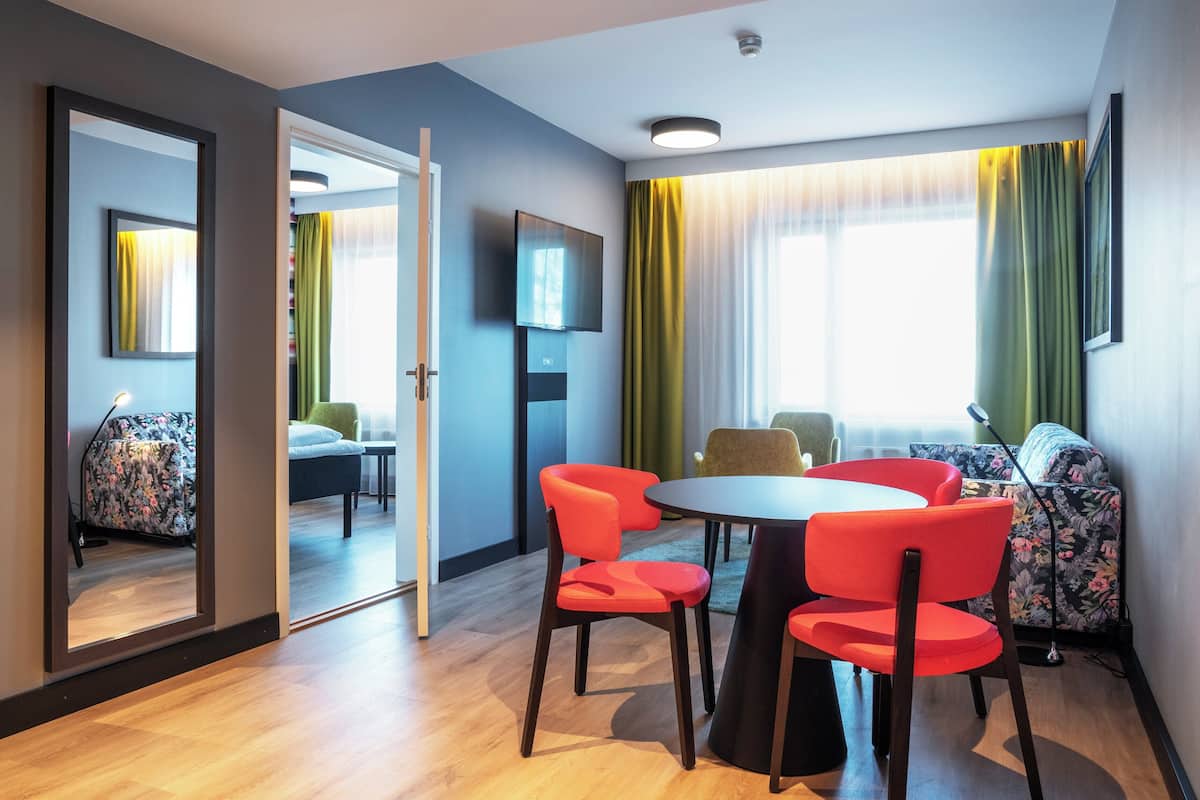 Een moderne hotelkamer met een ronde tafel, rode stoelen, een bank en een televisie aan de muur.