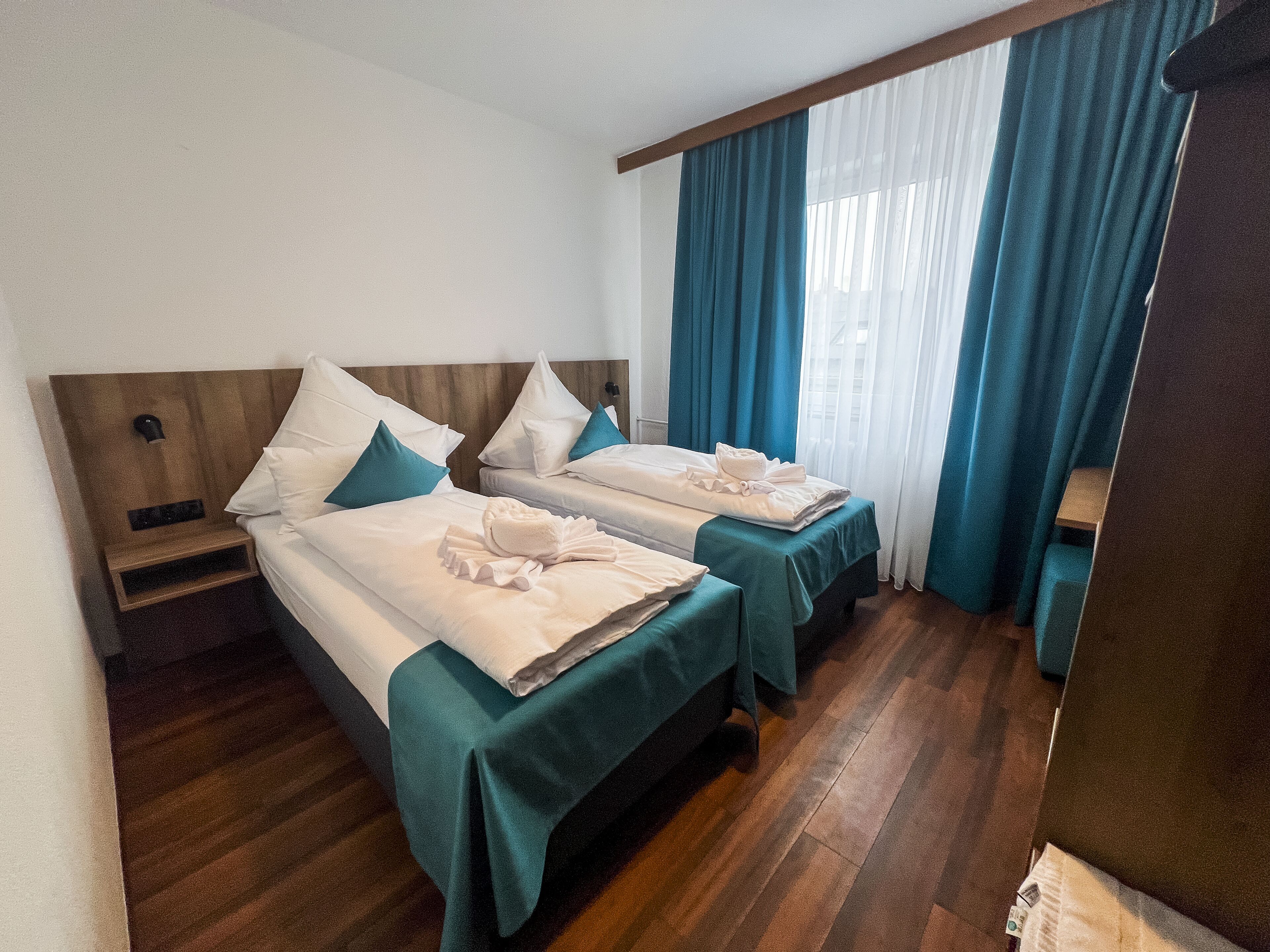 Ein Hotelzimmer mit zwei Betten, Holz-Kopfteilen, türkisblauen Bettdecken und weißen Bettwäsche, sowie einem Holzboden.