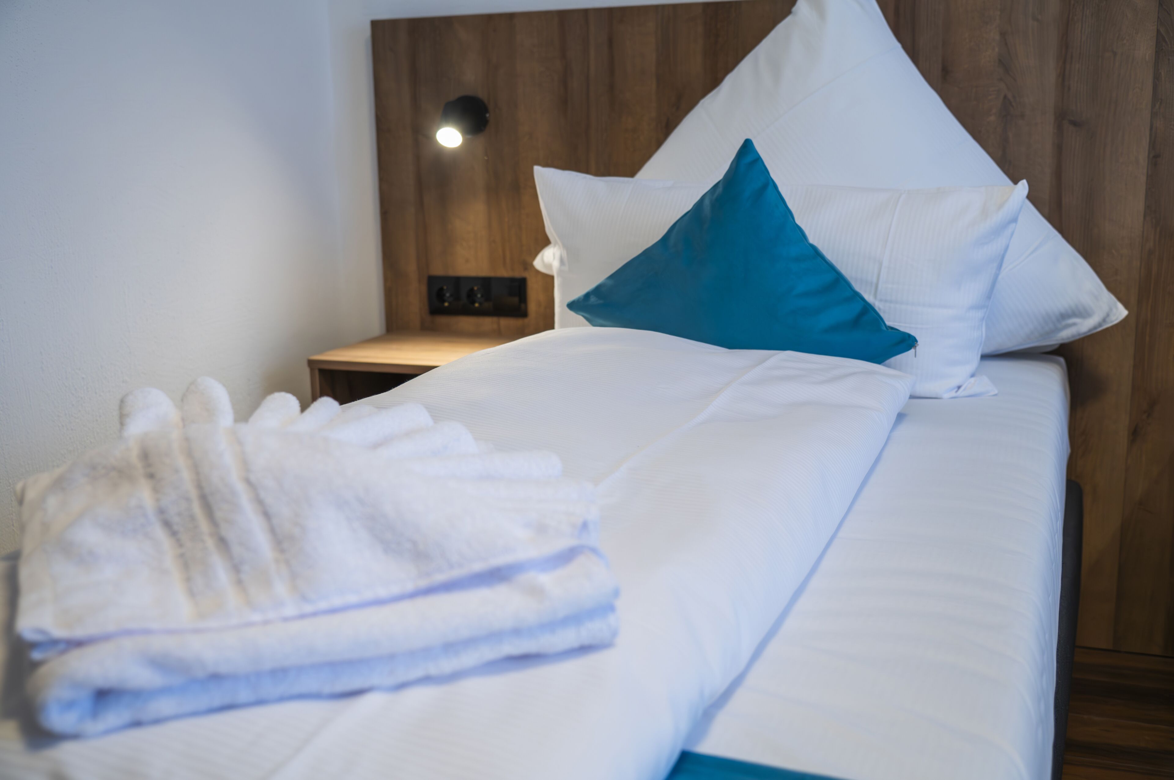 A neatly made bed with white and blue pillows, a folded white towel, and a wooden headboard with a wall-mounted light.
