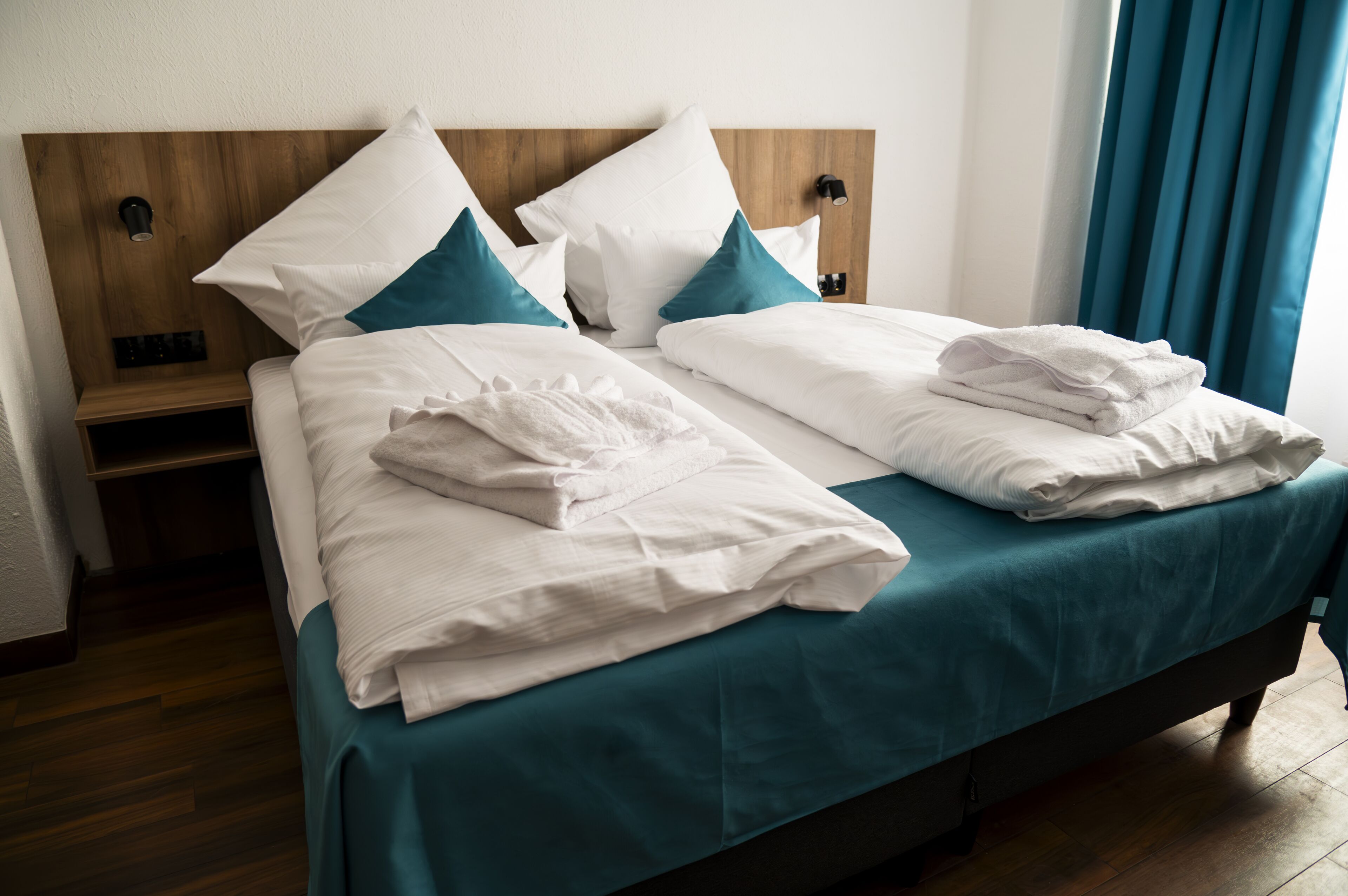 A double bed with white linens and teal bedding, a wooden headboard, and a nightstand with a lamp.