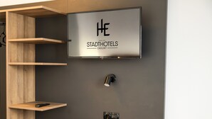 A modern hotel room with a mounted television displaying the logo of Stadthotels Erkurt, wooden shelves, and a small table. - Hotel Hansa (Stuttgart)