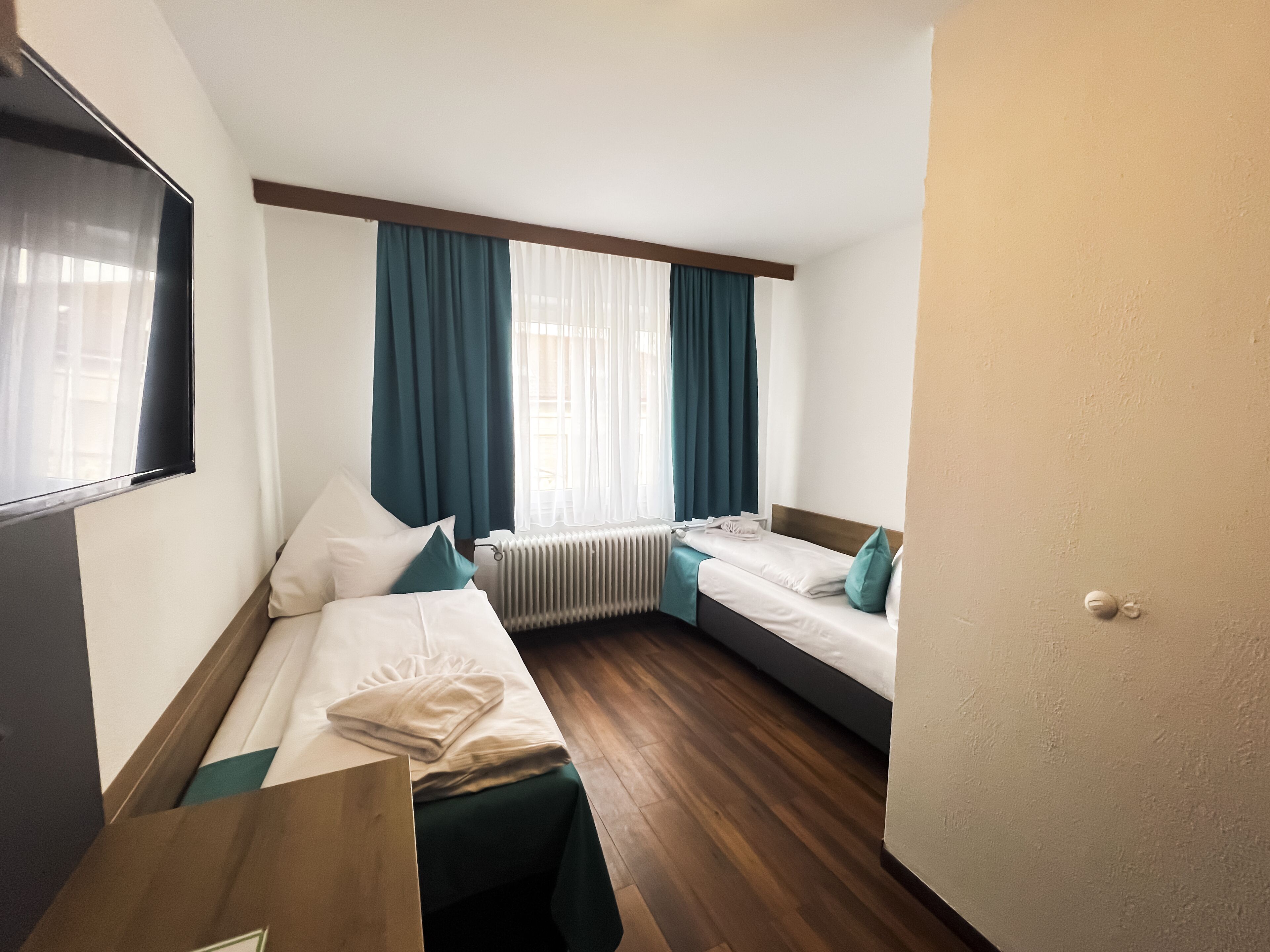 A hotel room with two single beds, a wooden floor, a large window with curtains, a radiator, and a TV mounted on the wall.