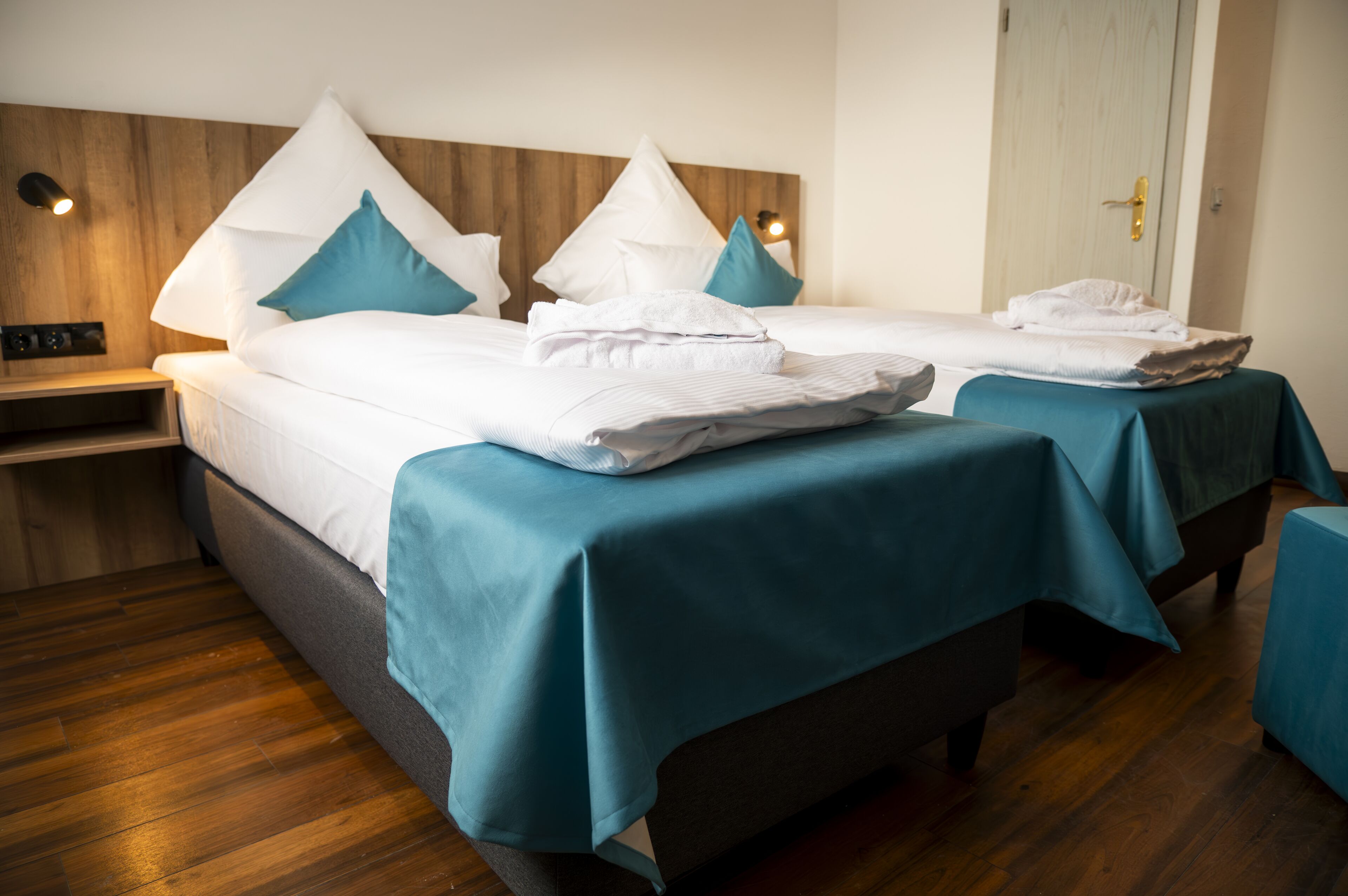 A hotel room with two single beds, each with a teal bedspread and white linens. There is a wooden headboard, a nightstand with a lamp, and a door with a golden handle.