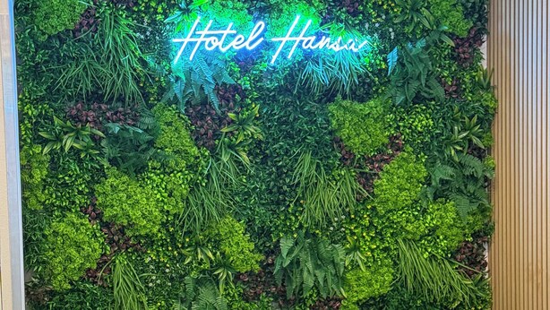 A black leather sofa with floral cushions, a round wooden coffee table with a floral vase, and a neon sign reading "Hotel Hans" against a backdrop of a lush green vertical garden. - Hotel Hansa (Stuttgart)