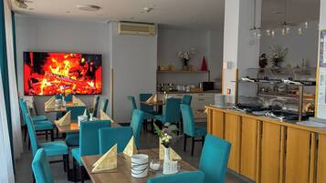 Restaurante com cadeiras cor de verde-água, área de buffet e televisão de tela plana exibindo uma lareira.