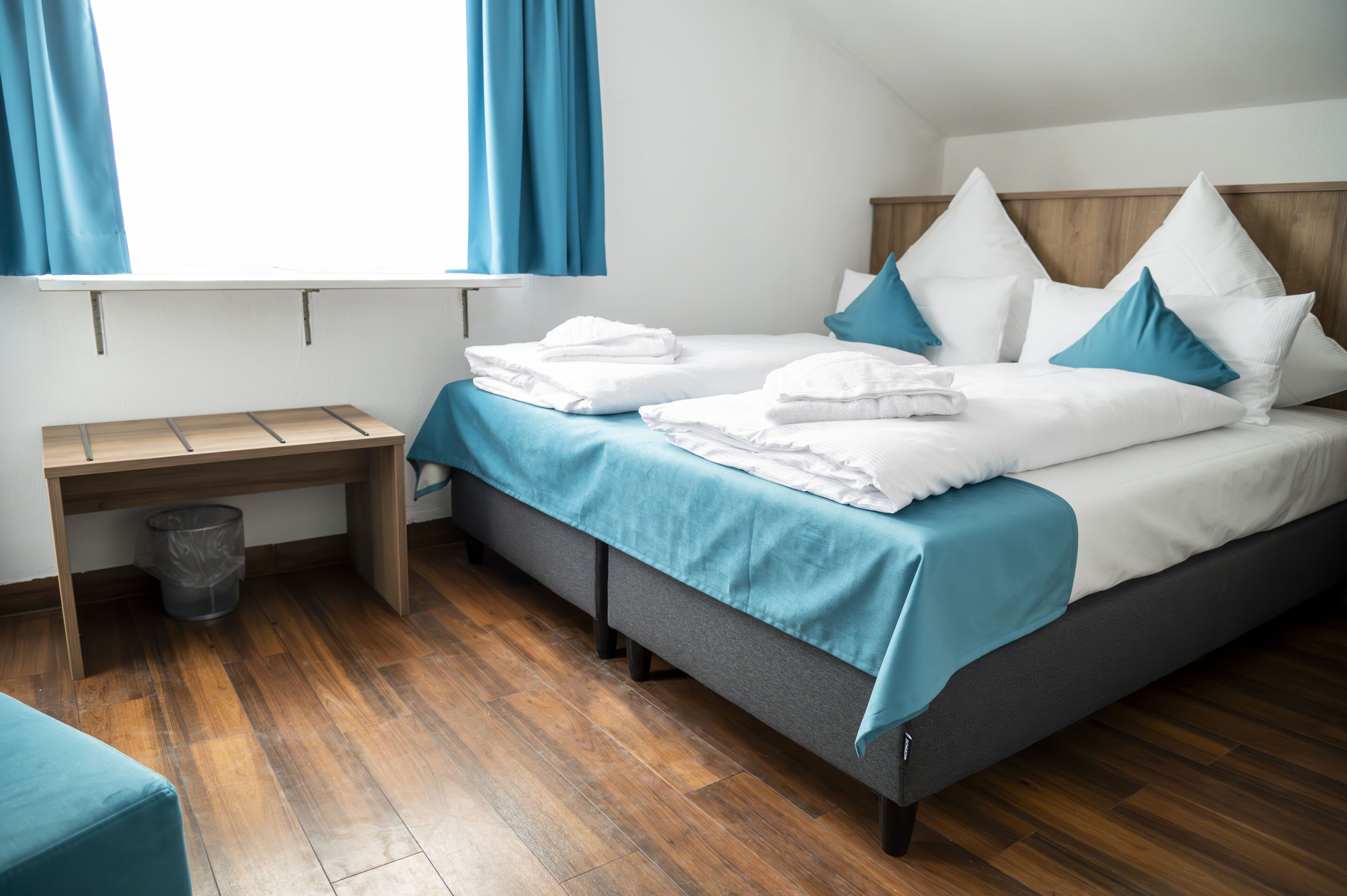 Una cama bien hecha con ropa de cama blanca y azul, un escritorio de madera con un cubo de basura y una ventana con cortinas azules.