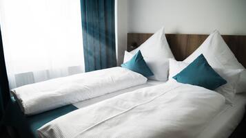 A hotel room with two beds, each with white bedding and teal pillows, a wooden headboard, and a window with sheer curtains.