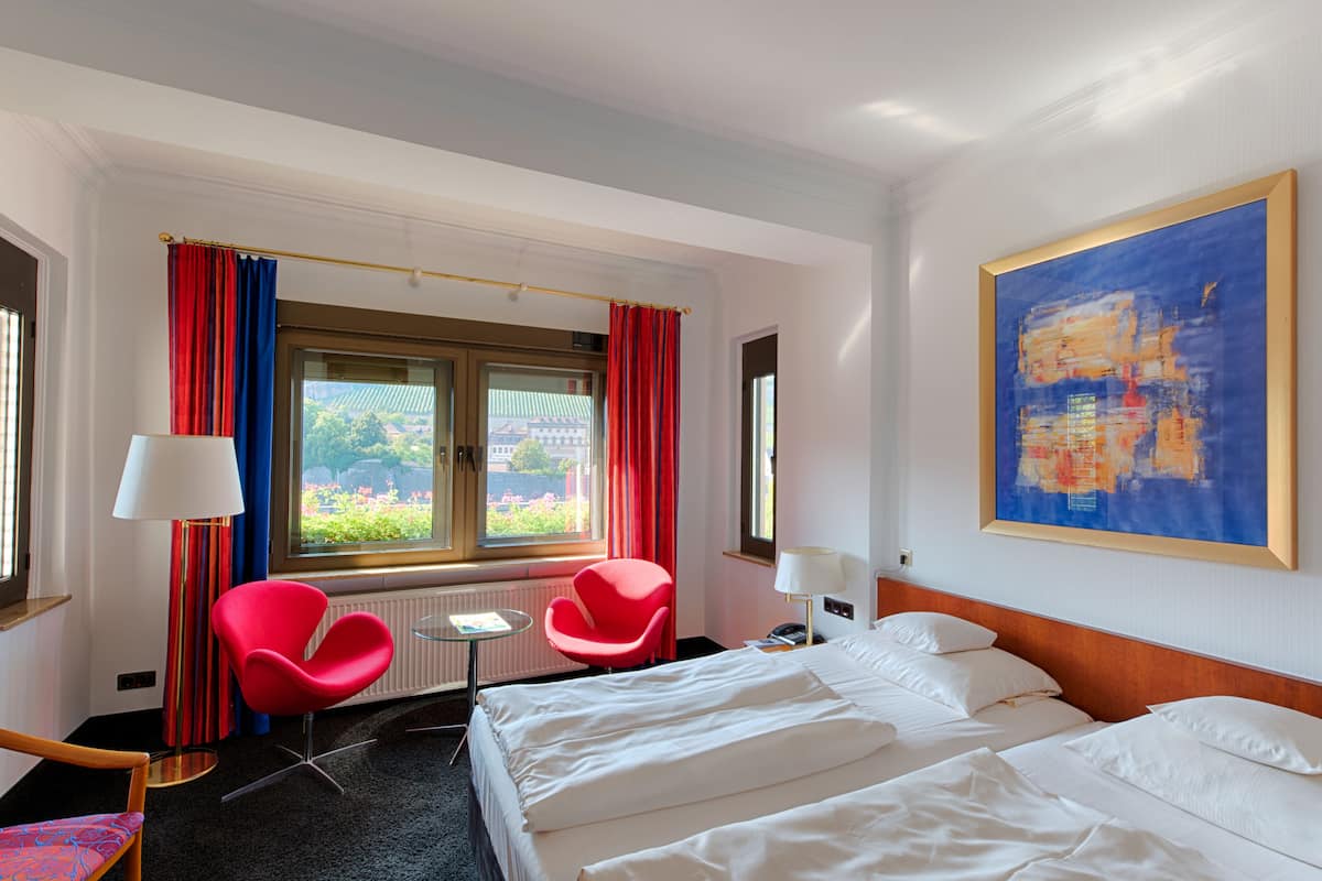 Una habitación de hotel con una cama, dos sillas rojas, una mesita pequeña, una ventana con cortinas rojas y un cuadro abstracto grande en la pared.