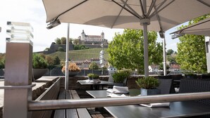 Een terras met tafels en parasols, met uitzicht op een heuvel met gebouwen en wijngaarden.