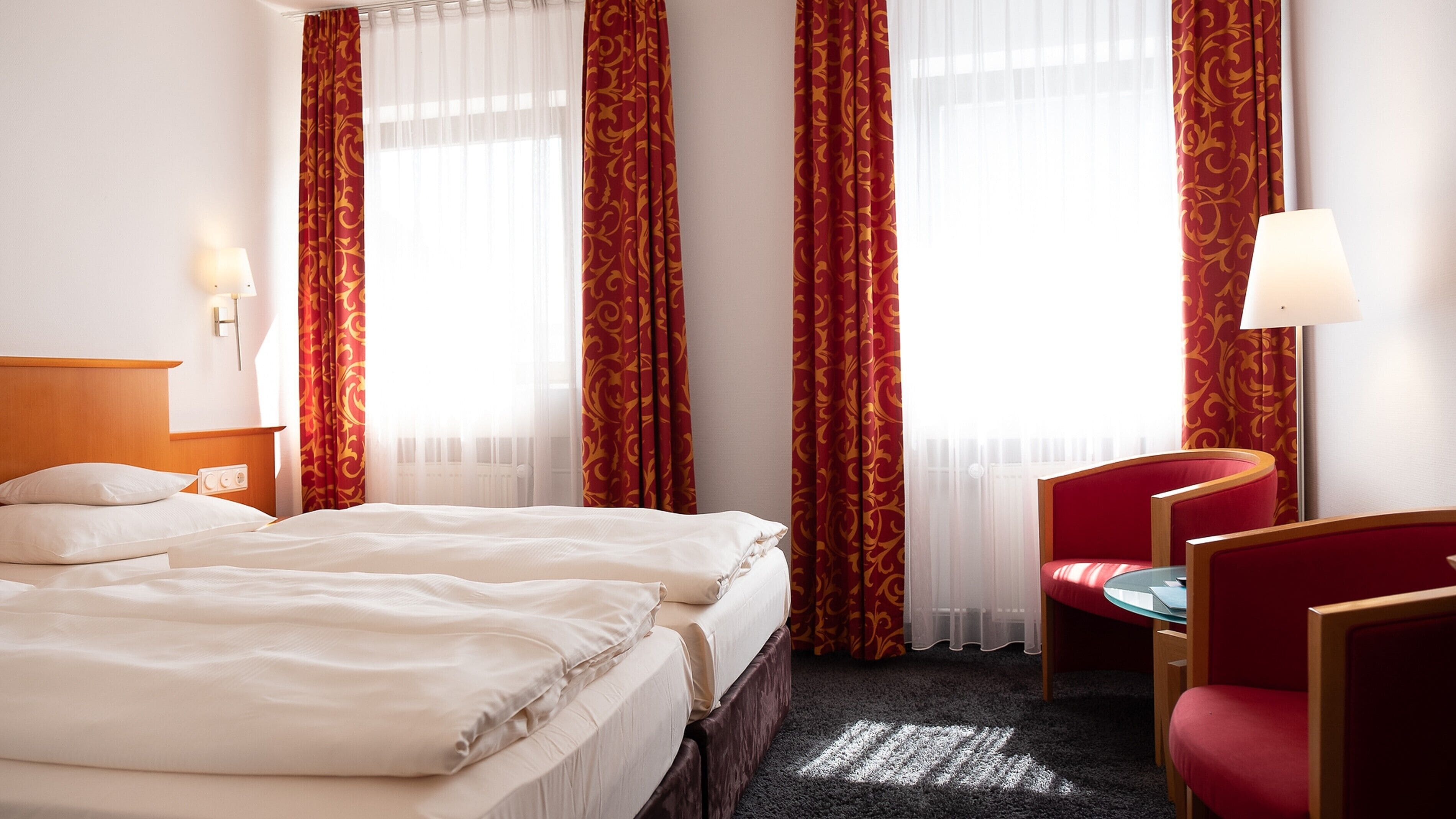 A hotel room with two beds, a desk, a chair, and a lamp.