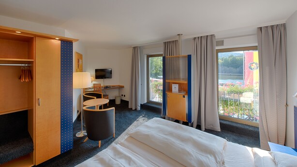 Een hotelkamer met een bed, bureau, stoel, televisie en uitzicht op een balkon met planten.