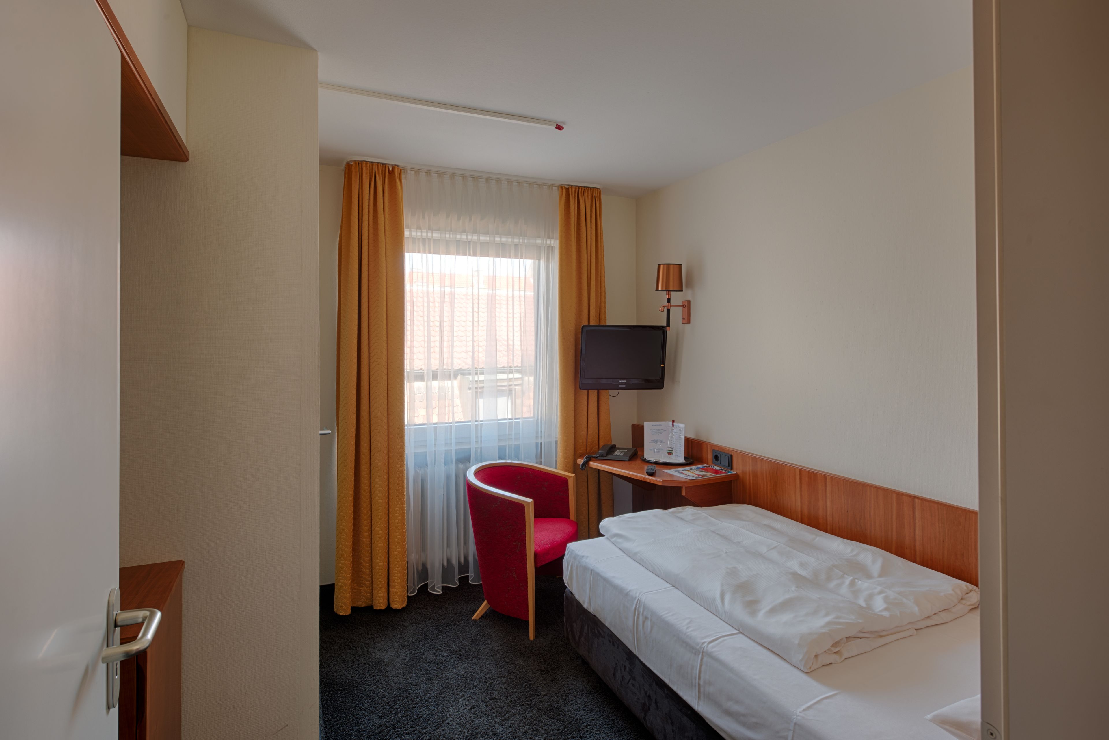 Une chambre d’hôtel avec un lit, un bureau avec un ordinateur, une chaise, une télévision et une fenêtre avec des rideaux.