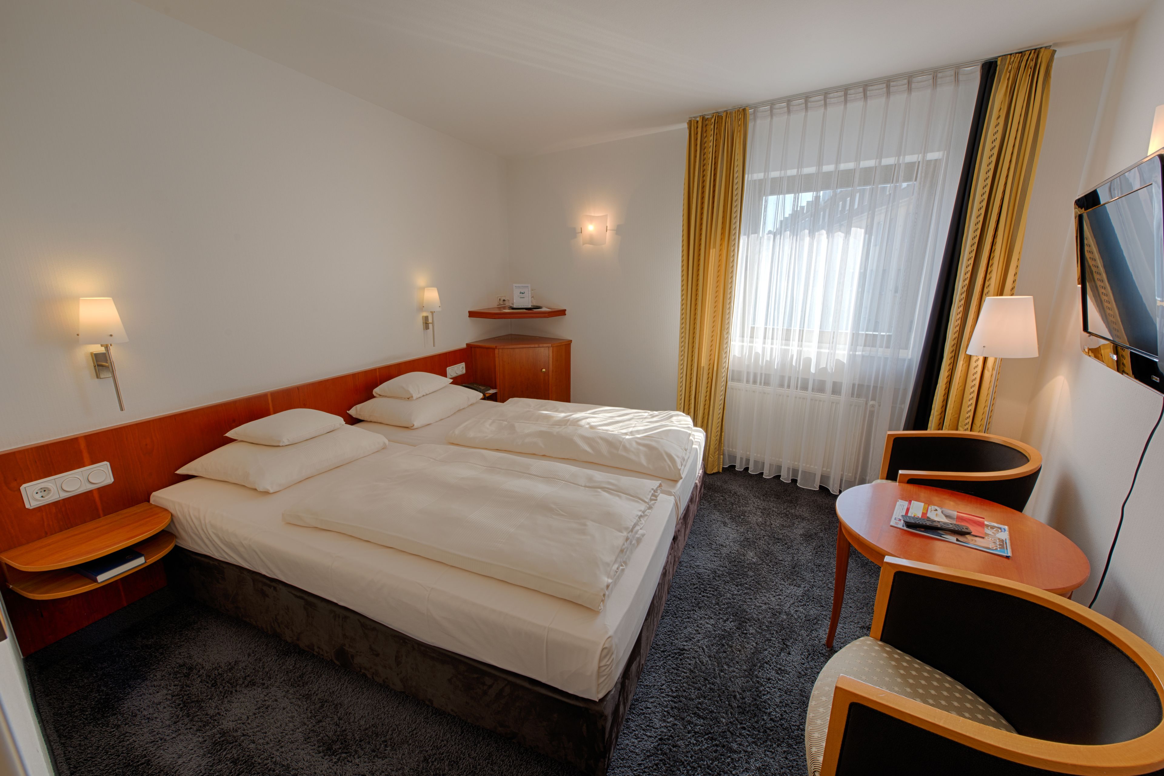 Ein Hotelzimmer mit einem großen Bett, Nachttischen, einem Schreibtisch mit Stuhl, einem Fernseher und einem Fenster mit Vorhängen.