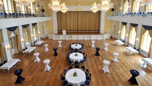 Een ruime balzaal met meerdere ronde tafels, hoge plafonds en versierde kroonluchters.