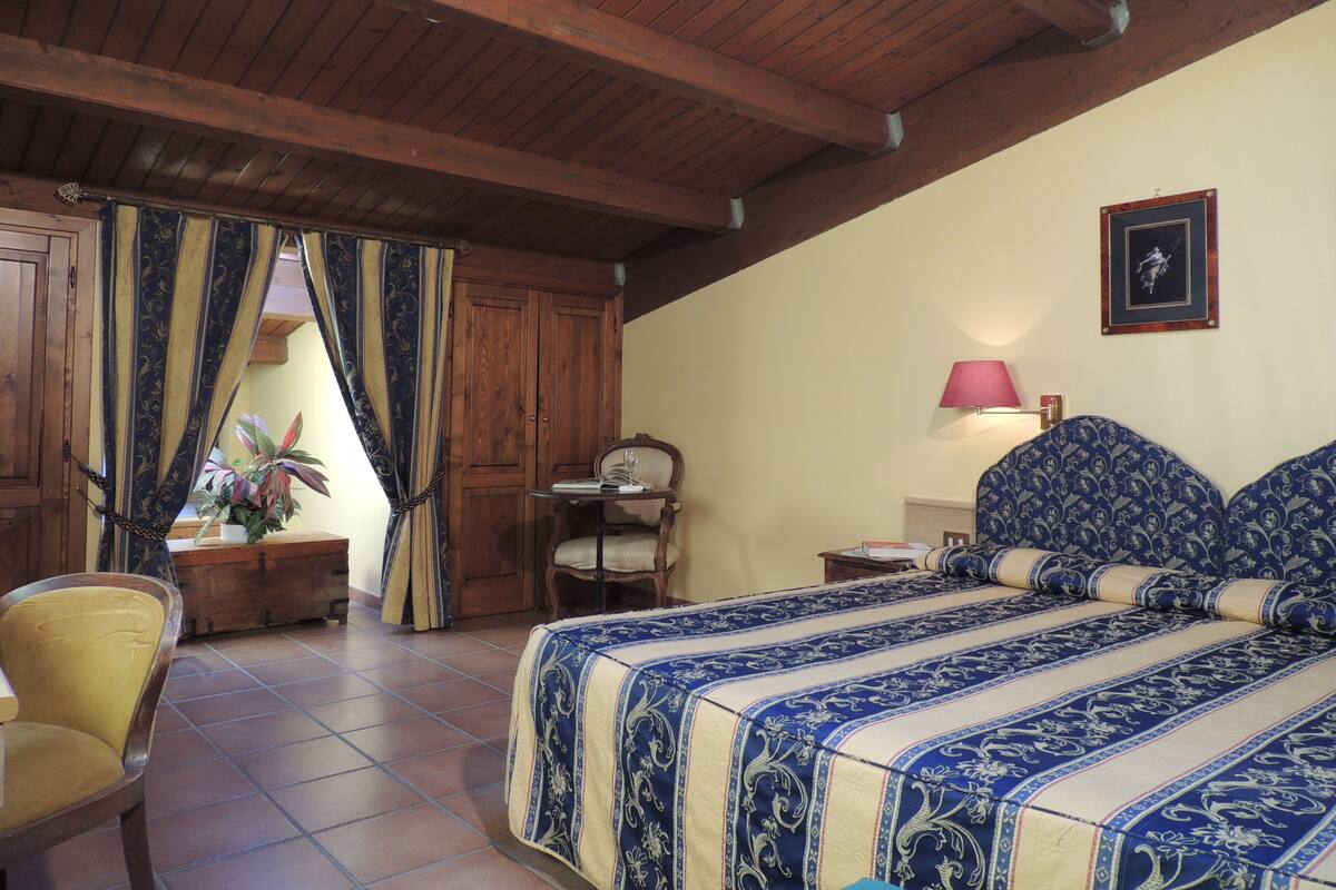 A bedroom with a wooden ceiling, a bed with a blue and white patterned bedspread, a bedside table with a lamp, a chair, and a wooden wardrobe.