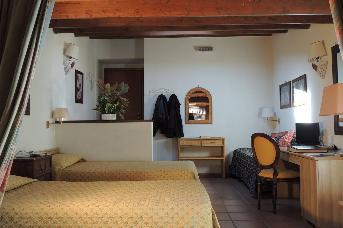 A hotel room with two beds, a desk, a chair, a small table, a lamp, a plant, and a mirror.