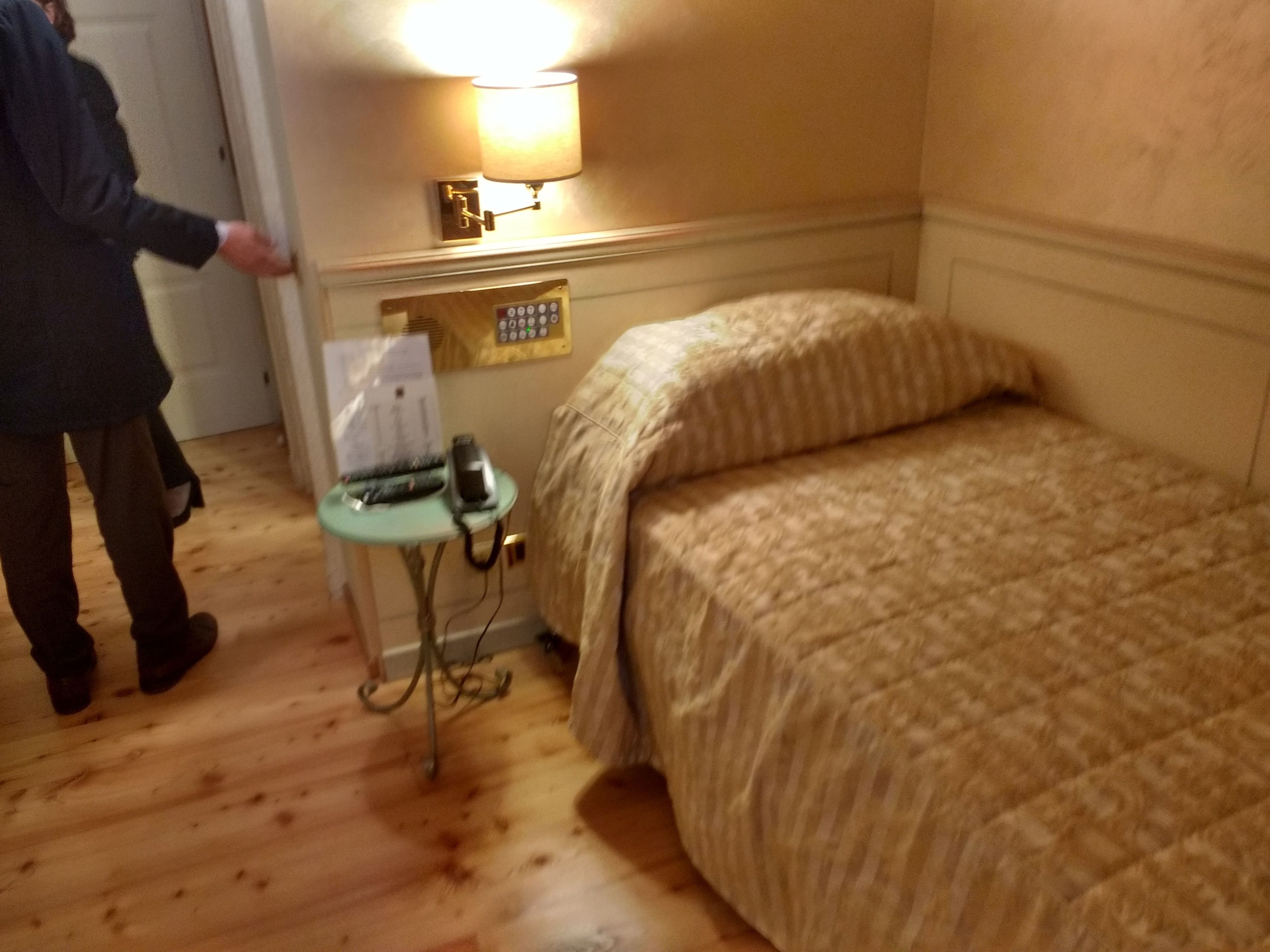 A hotel room with a wooden floor, a bed with a striped blanket, a bedside table with a lamp, a telephone, and a wall-mounted control panel.