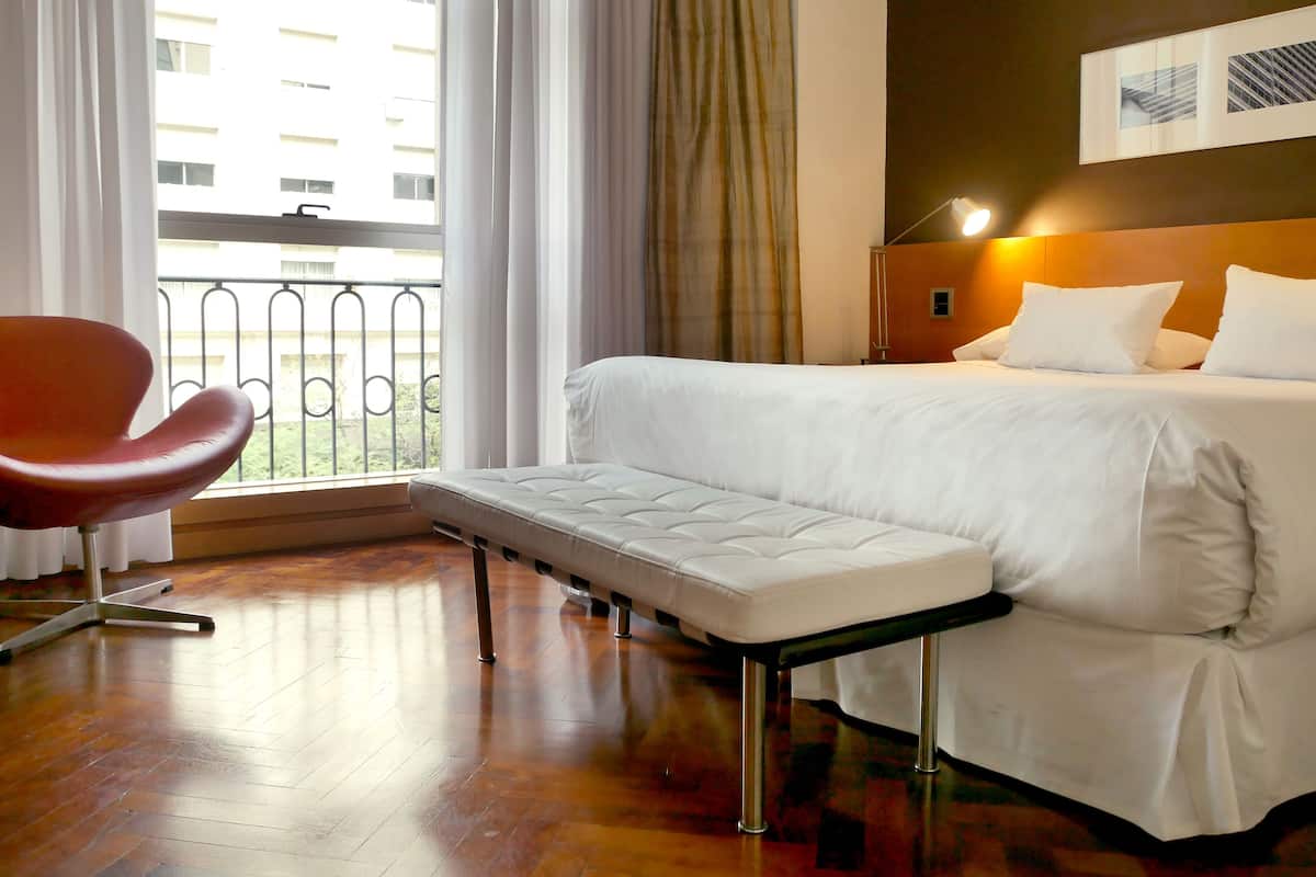 A hotel room with a bed, a bench, a chair, a window with curtains, and a view of a building outside.