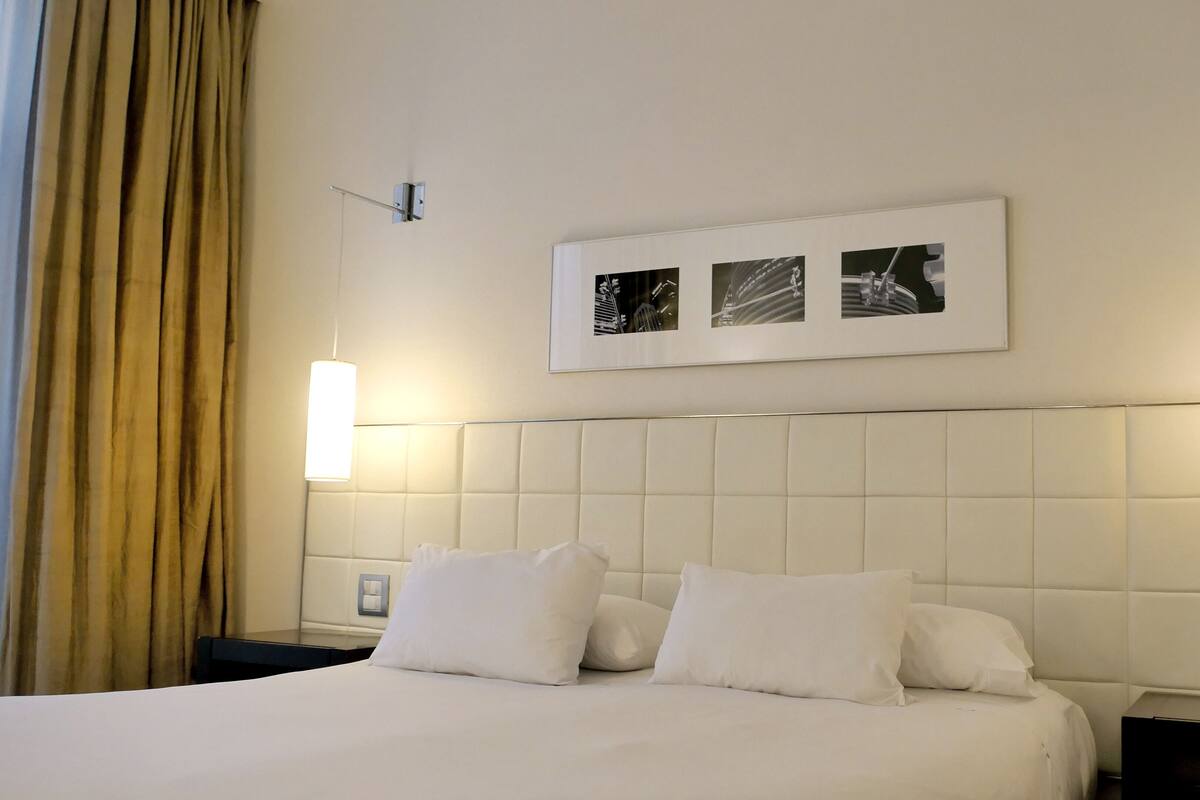 A hotel room with a large bed, two bedside lamps, a headboard with a picture, and a window with curtains.