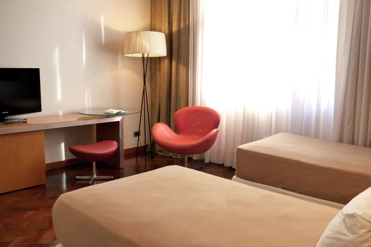 A hotel room with a TV, a desk, a red chair, two beds, and a lamp.