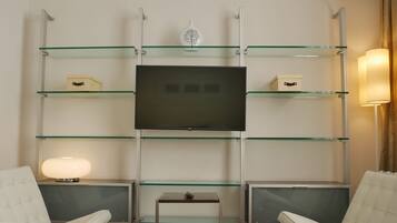Ett modernt rum med en platt-TV som är monterad på en hylla av glas, två vita fåtöljer med vaddering, ett litet bord och en sänglampa.