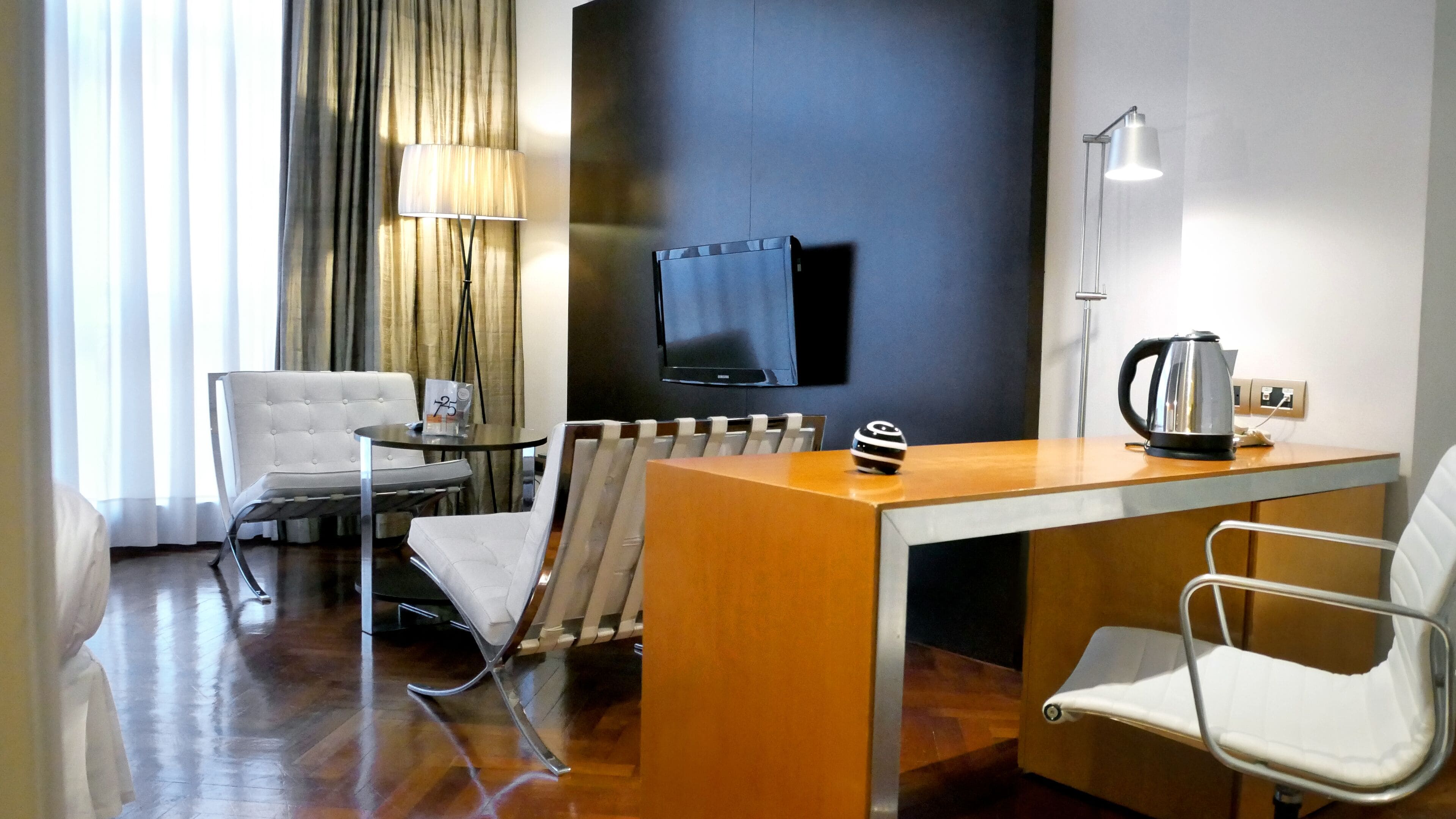 Habitación de hotel con un escritorio de madera, dos sillas blancas, un televisor, una lámpara y un hervidor.