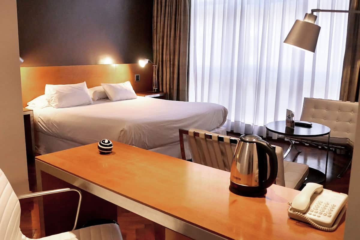 A hotel room with a bed, a desk with a kettle, a chair, a table, a lamp, and a telephone.