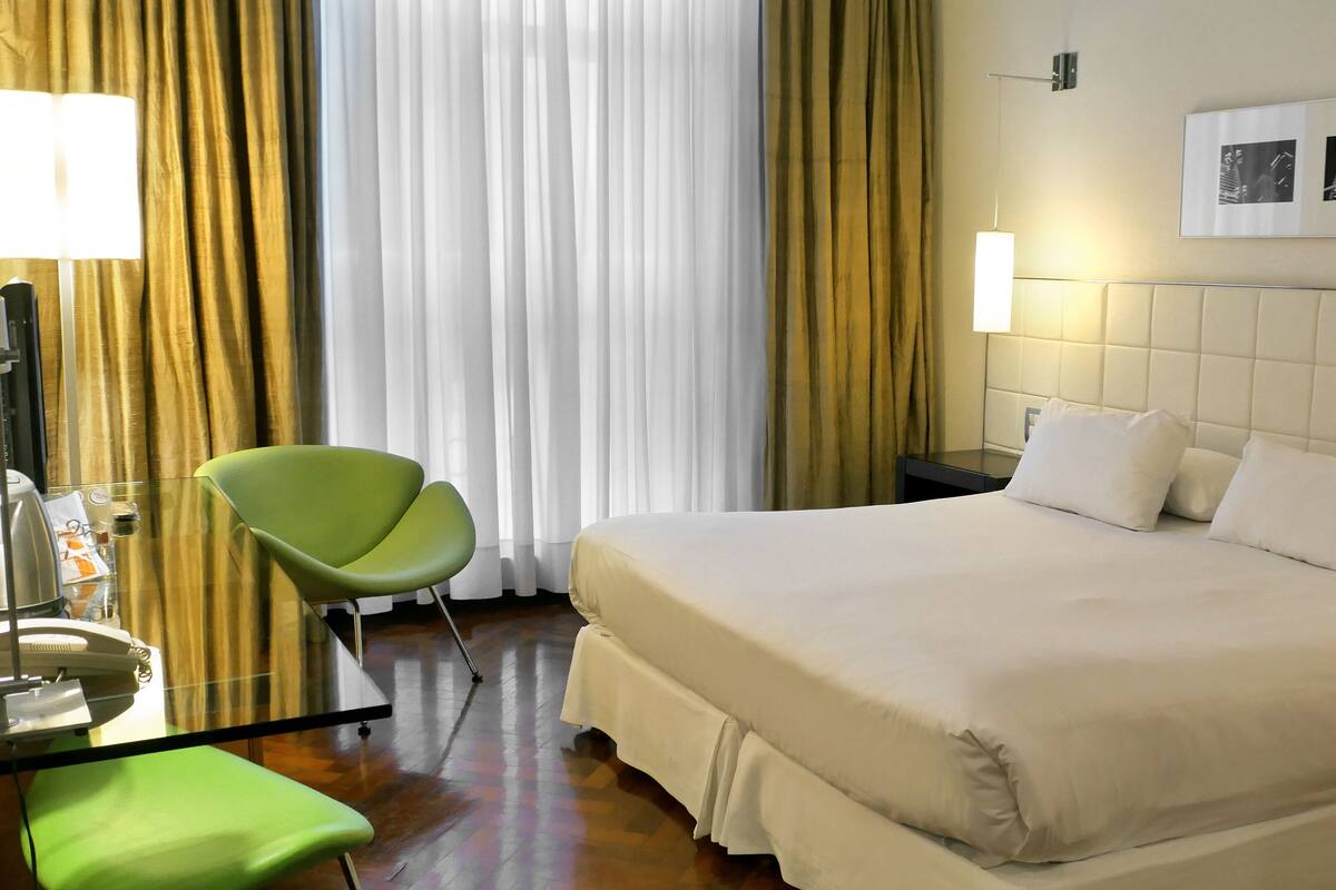 A hotel room with a large bed, a green chair, a bedside table, and a lamp.