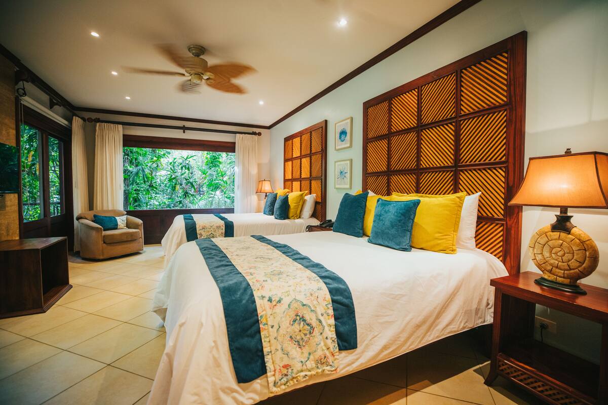 A hotel room with two beds, a ceiling fan, a window with curtains, a wooden partition, and a lamp on a bedside table.