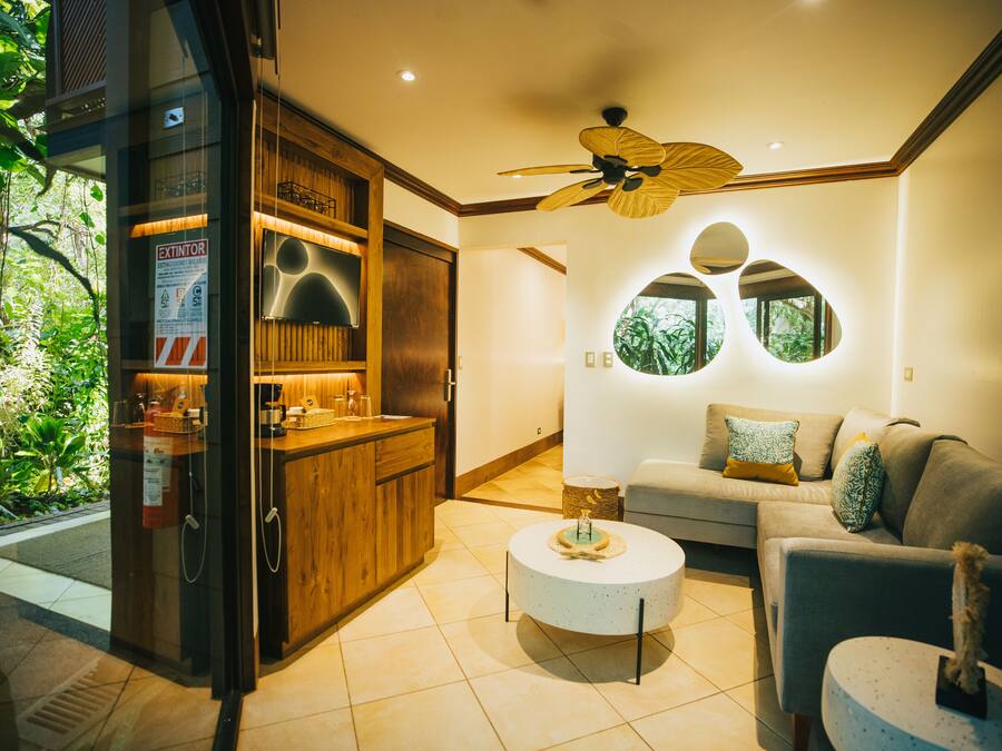 A modern living room with a sofa, coffee table, and a ceiling fan. There is a kitchen area with wooden cabinets and a glass door leading outside.