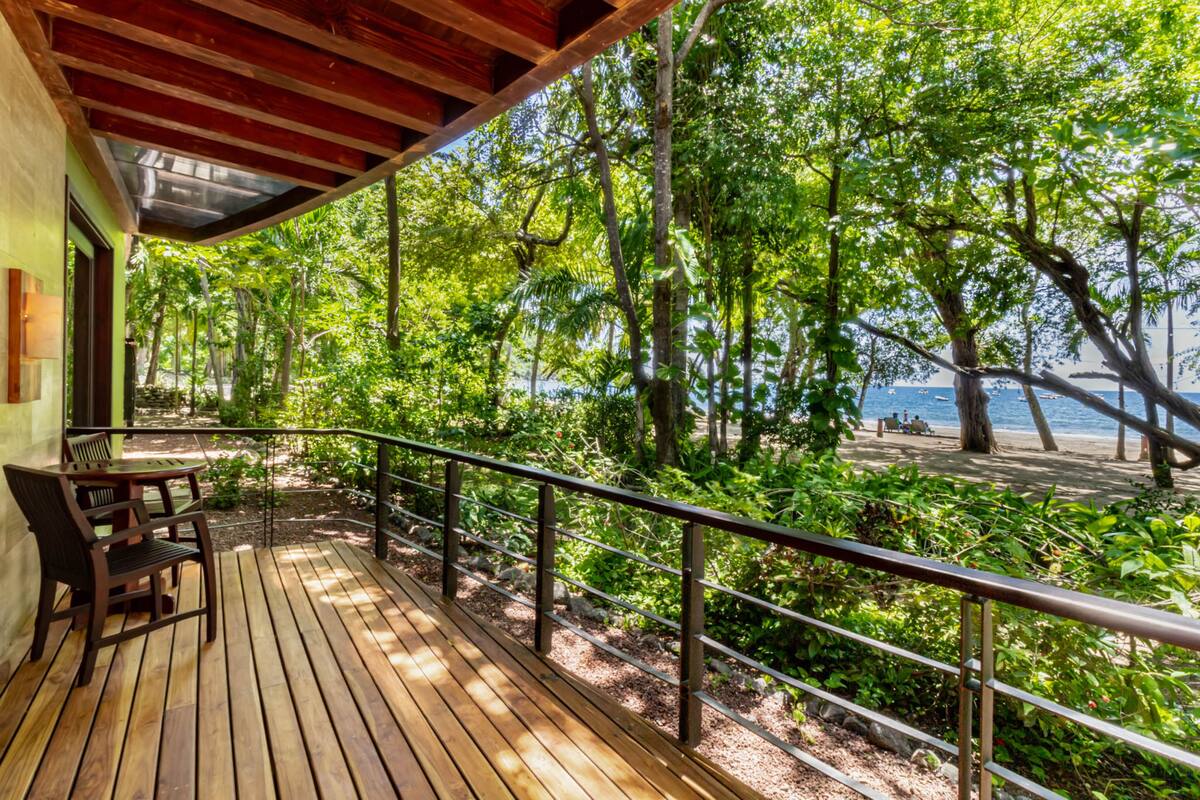 A wooden deck with a metal railing overlooking a beach with trees and a clear view of the ocean.