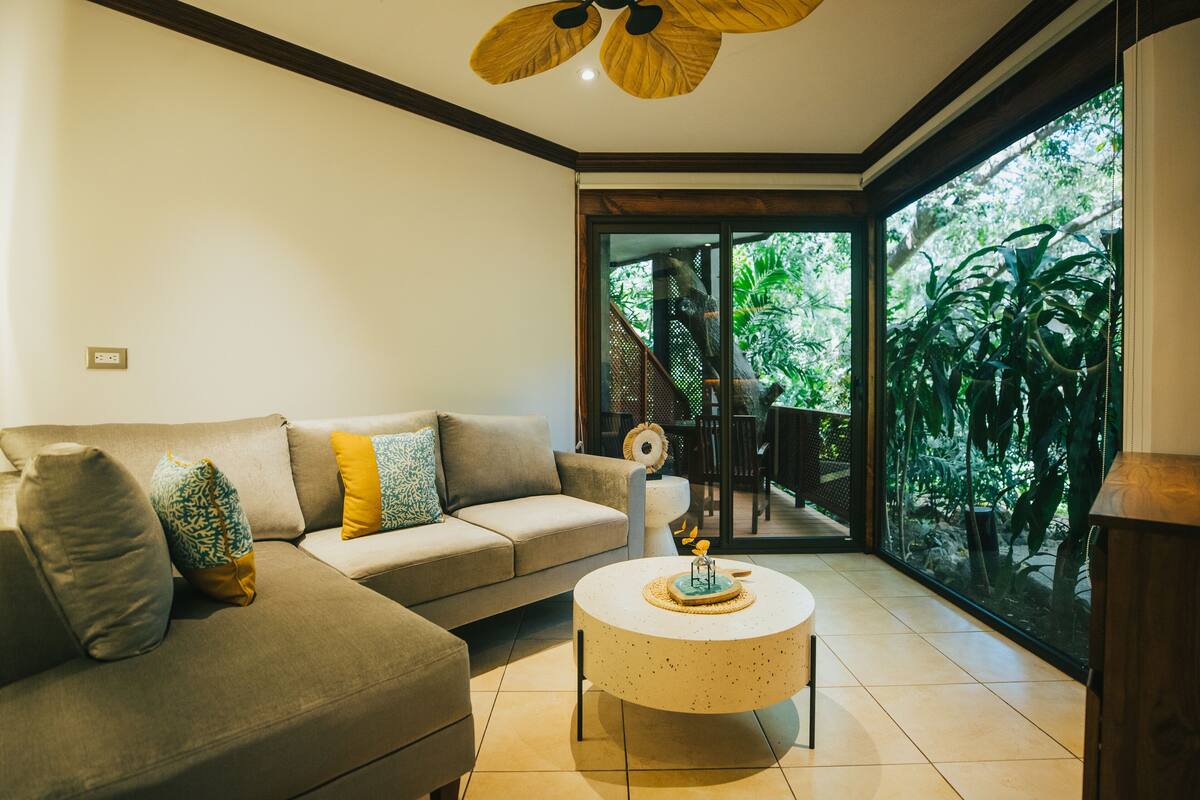 A modern living room with a beige sectional sofa, a round coffee table, and a large glass door leading to a balcony with greenery.