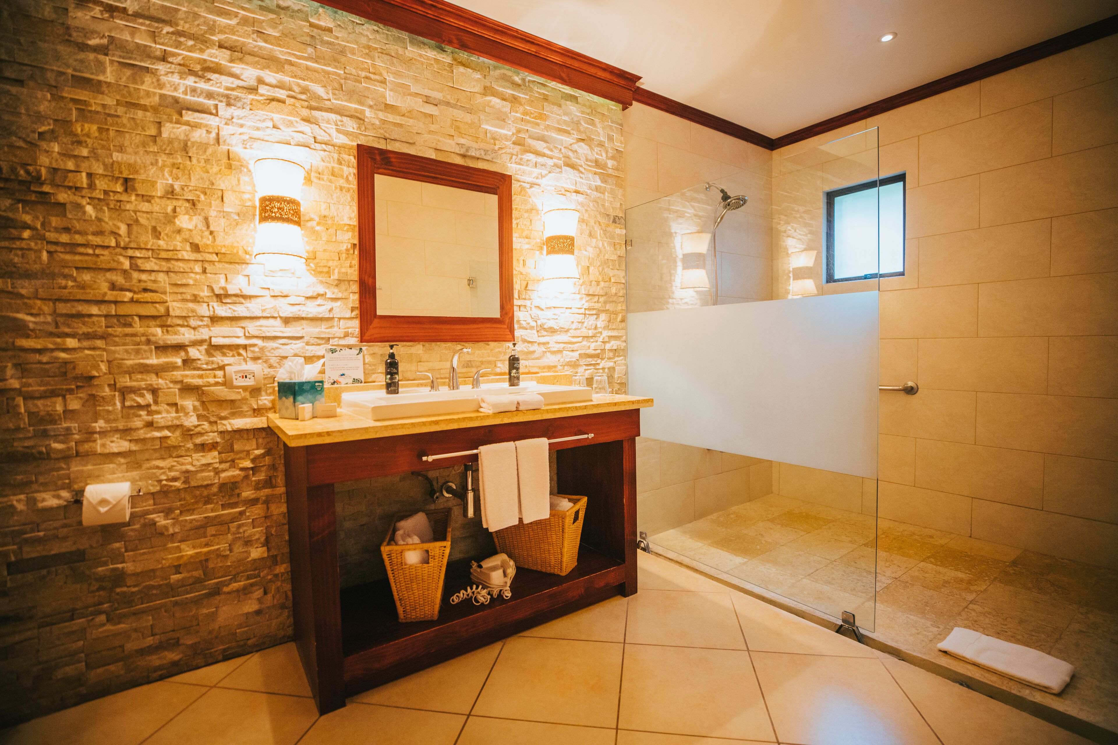 Un baño con pared de piedra, tocador de madera, cabina de ducha de vidrio y espejo sobre el lavabo.