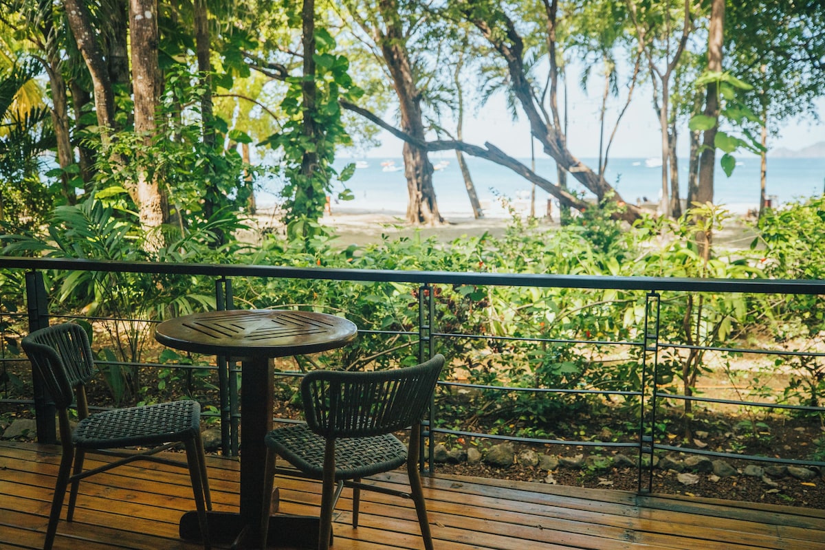 A wooden deck with two wicker chairs and a table, overlooking a beach with trees and a clear sky.