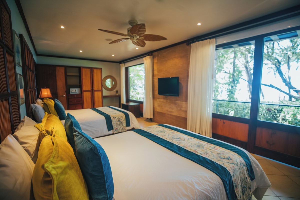 A hotel room with two beds, a ceiling fan, a television, and a large window with a view of trees.