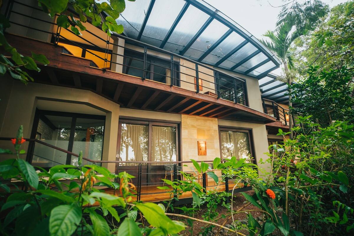 A modern two-story house with a glass roof, wooden balconies, and a garden in the foreground.